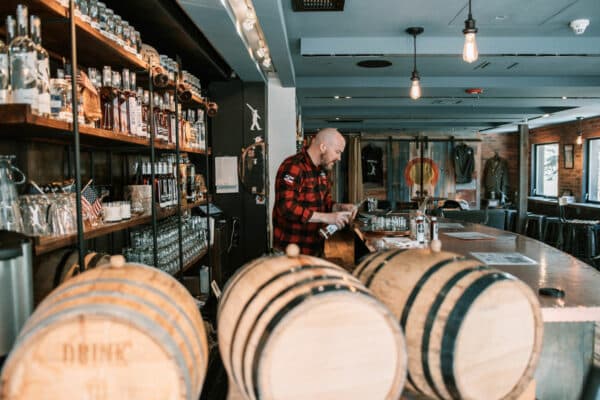 A man wearing a black-and-red plaid shirt tends to the bar at 10th Mountain Whiskey & Spirits Co. in Vail, Colorado. Wooden barrels sit in the foreground.