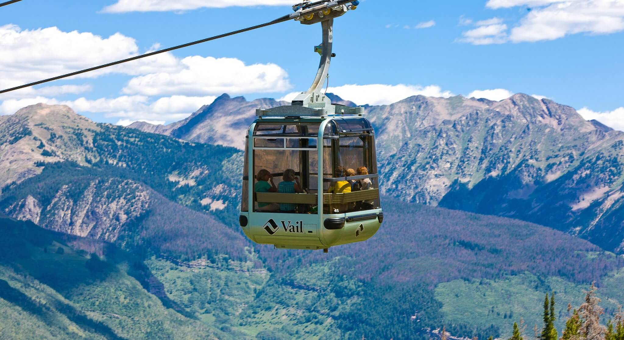 A silver gondola with a Vail logo carries people up the mountain on a cable, with lush, green mountains rising in the background under a blue sky.