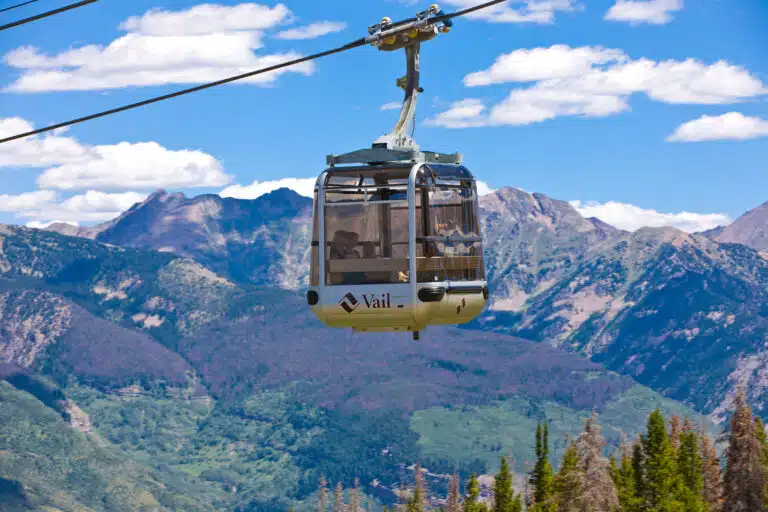 A silver gondola with a Vail logo on it heads up the mountain on a series of cables. Green trees and foliage blanket the terrain in the background, as does a grey-and-purple mountain ridge.