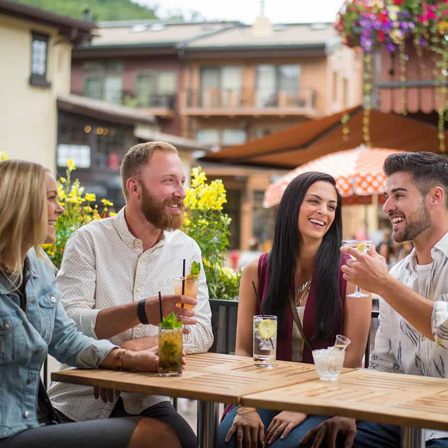 A smiling friend group of four sits at at an outdoor table at a Vail restaurant and chats. The Bavarian-style village stands behind them, with a robust flower arrangement dripping off a second-floor deck above them.