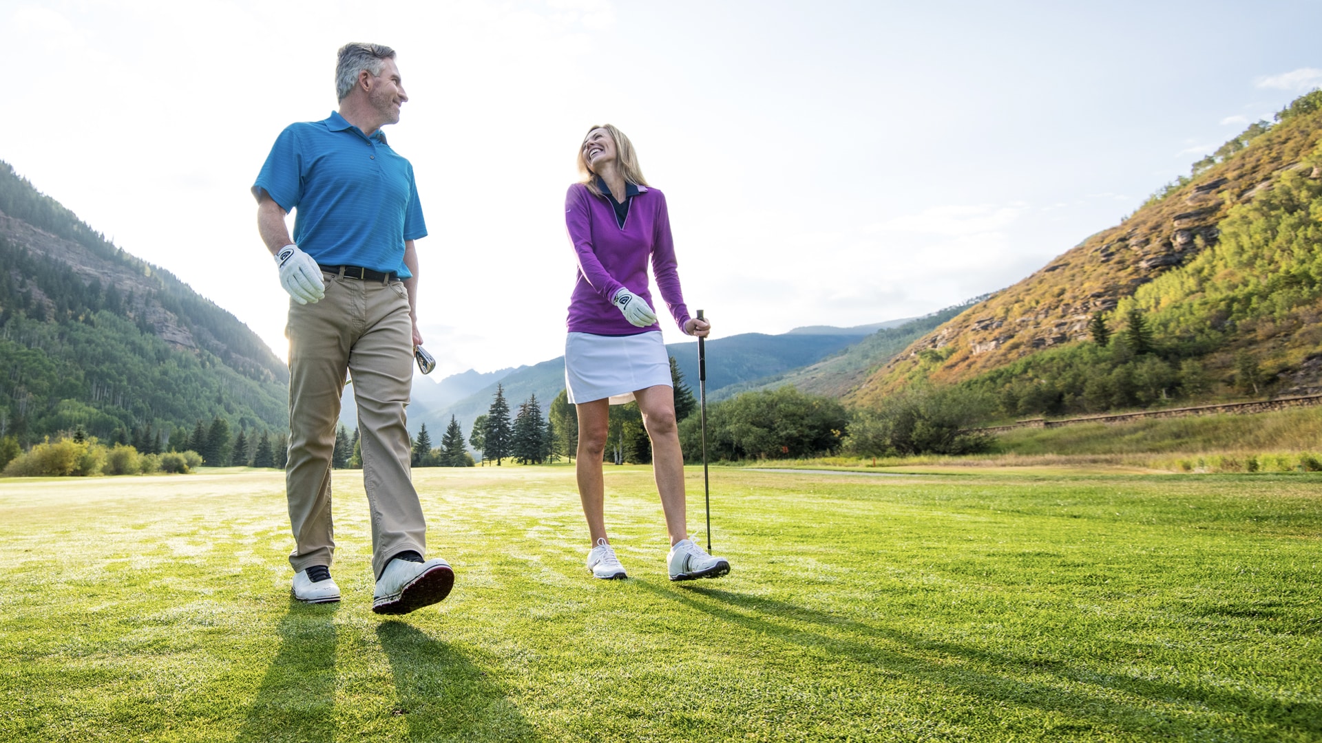 A man and woman walking on a golf course with mountains in the background.