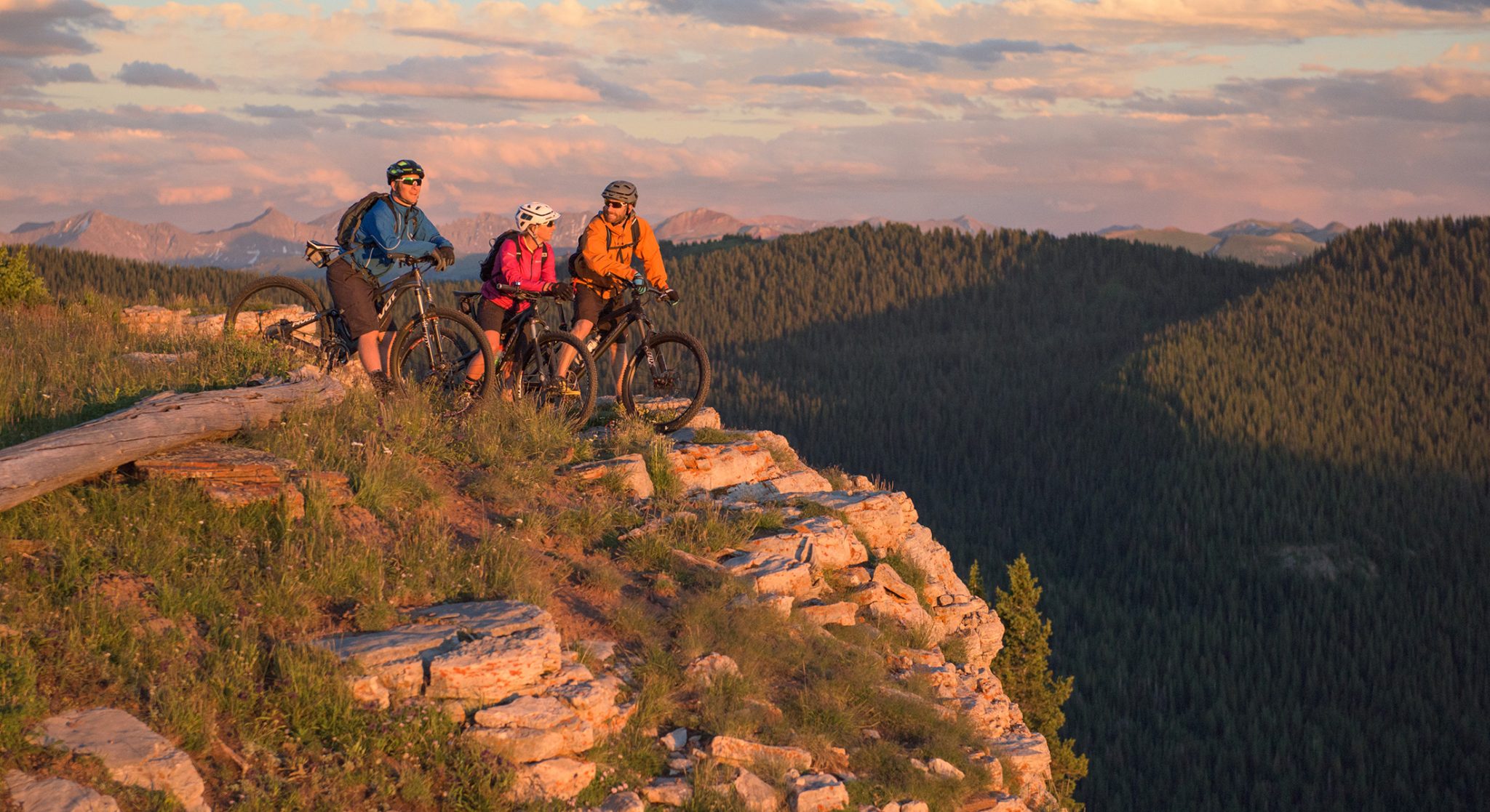Three helmeted mountain bikers in colorful jackets pause on a rocky outcropping to chat with views of the Rocky Mountains and evergreen trees in the background.