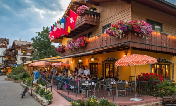 People relax at tables on a bar's patio in Vail Village. American and European flags line the balcony of the Bavarian-style restaurant above them. Boxes and bushes full of flowers of all colors surround the scene.