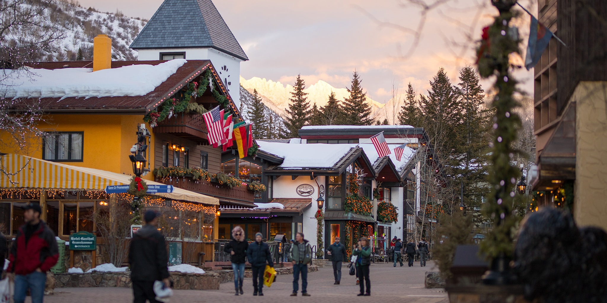 Several people stroll the brick-lined walkways of Vail Village at dusk. Several of the Bavarian-inspired building house shops and have snow-covered roofs.