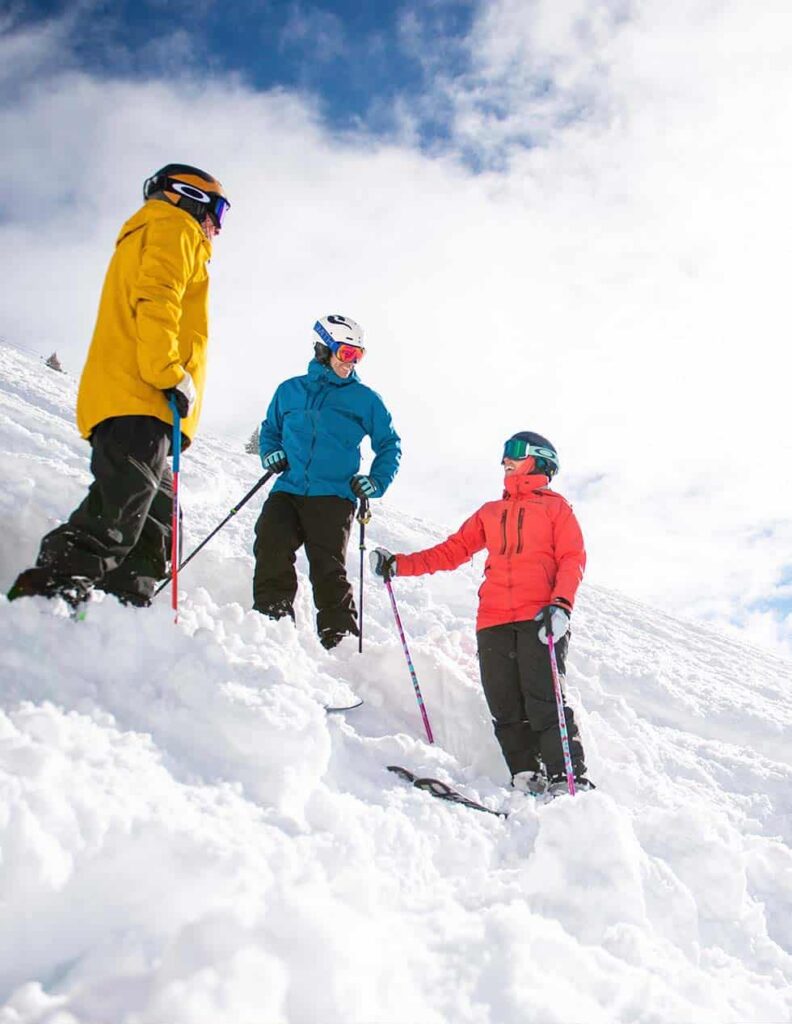 Three skiers in helmets, goggles and brightly colored coats take a break amid fresh, piled up snow on the side of Vail Mountain.
