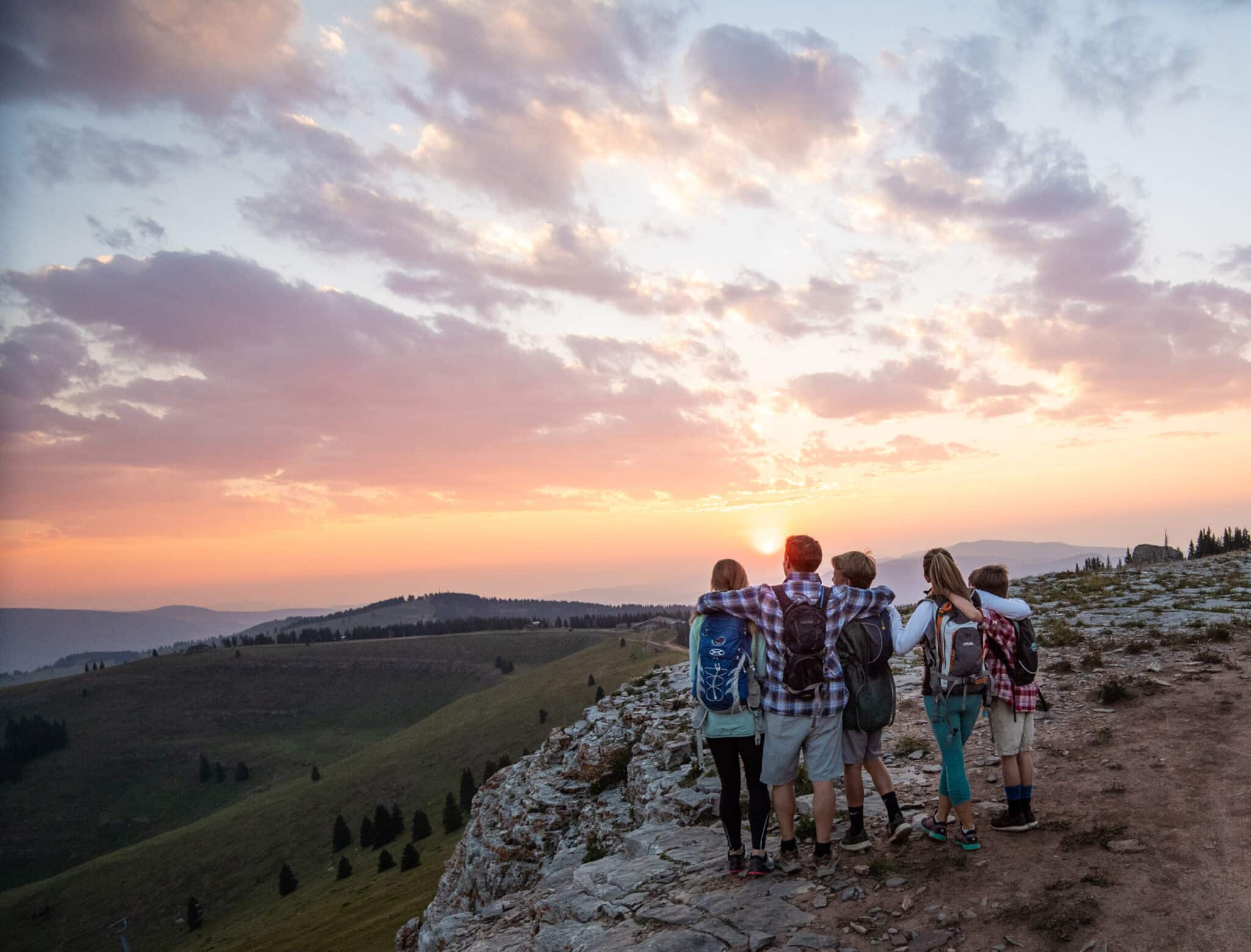 A family of five stands close together with their arms around each other as they watch a blazing sunset on a rocky outcropping in Vail, Colorado.