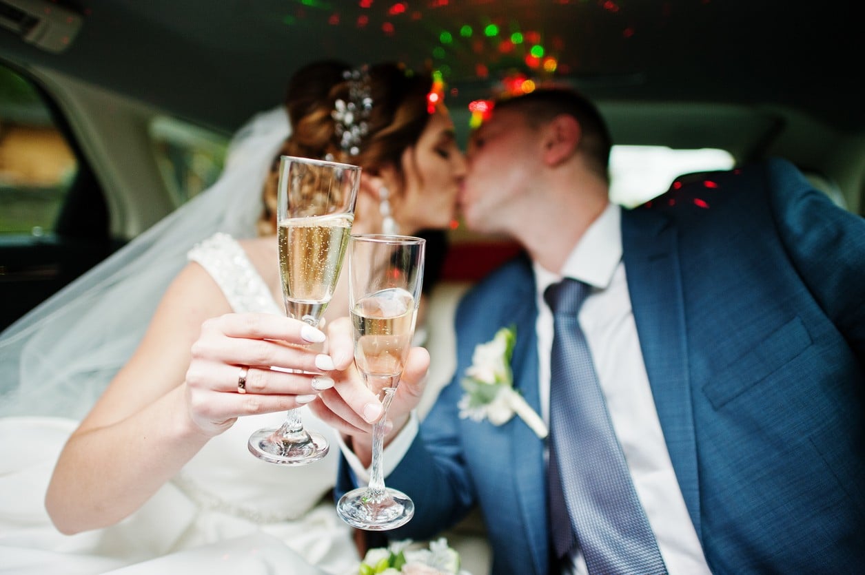 Bride and groom toasting and kissing in limo. Glasses of Champagne are in the foreground as they clink them in celebration.