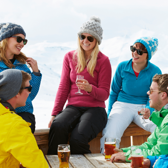 Group of smiling people wearing sunglasses and warm-looking hats enjoying apres-ski drinks in Vail, Colorado.