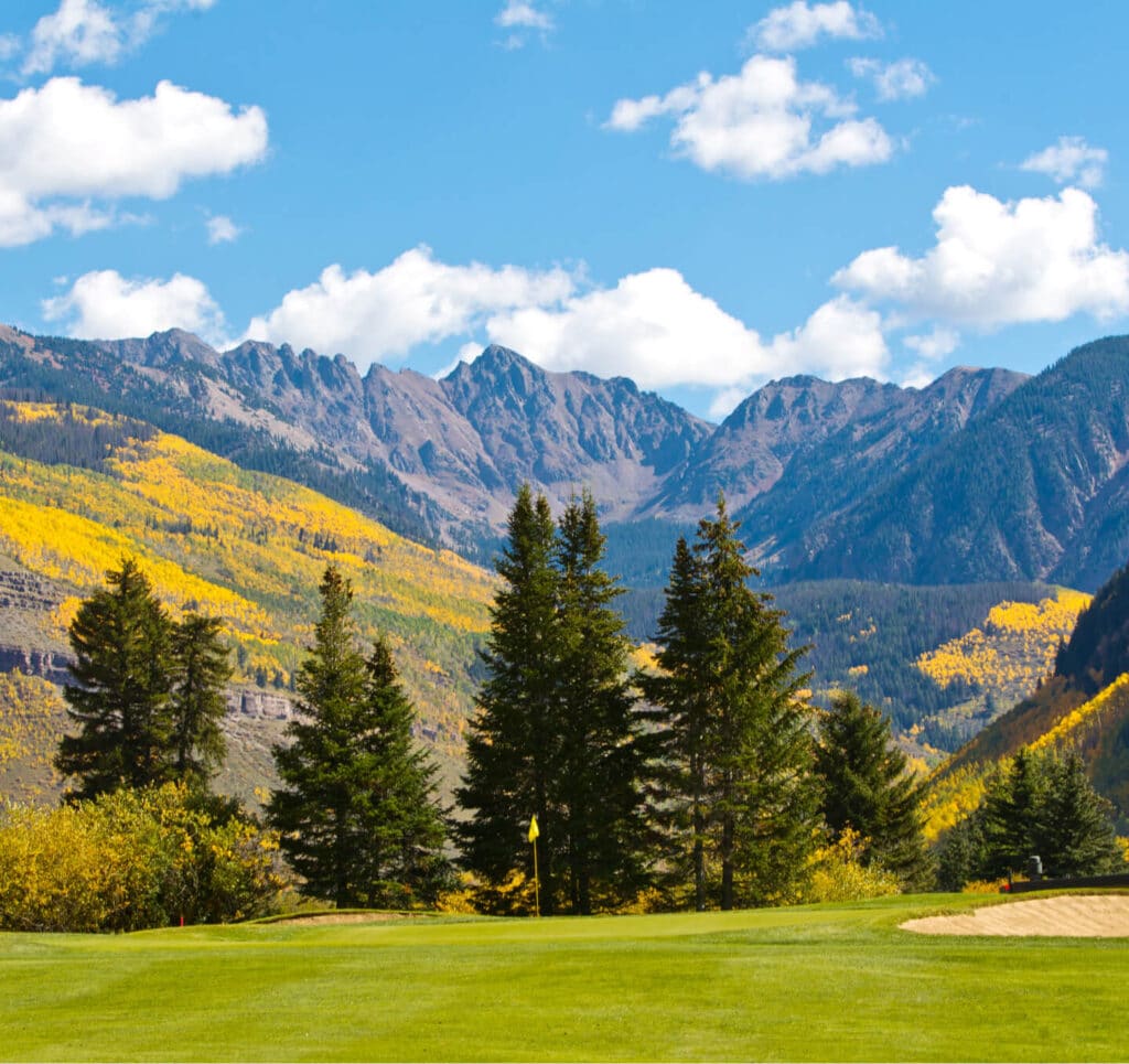 A green golf course in front of a line of evergreen trees, a purple mountain ridge and a mountainside covered by yellowing shrubs.