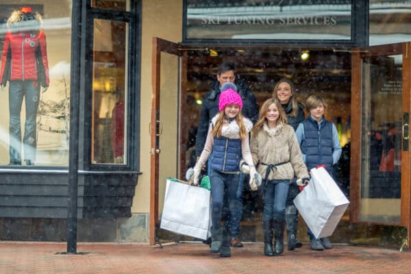 A family of five are out shopping in the Vail village during winter. They are leaving a store and two girls carry white shopping bags.