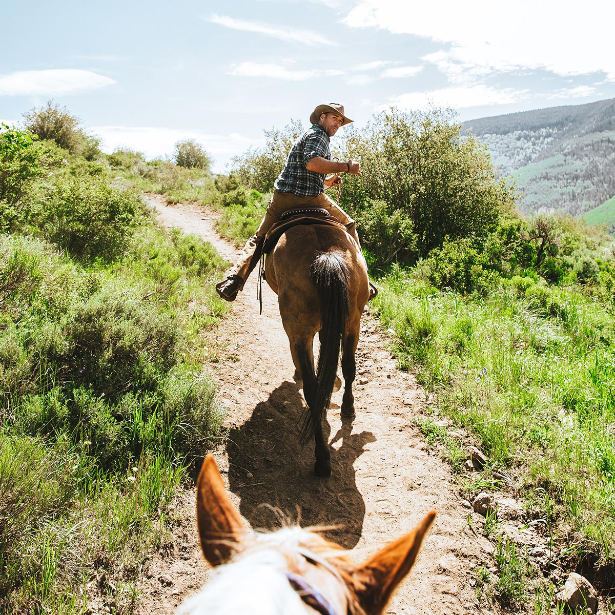 A person on a horse looks behind them at another horse riding the trail in Vail. The trail is surrounded by green grasses.