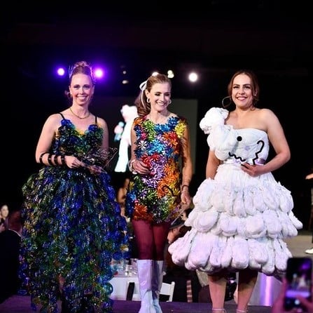 Three women standing on a stage wearing dresses made out of plastic.