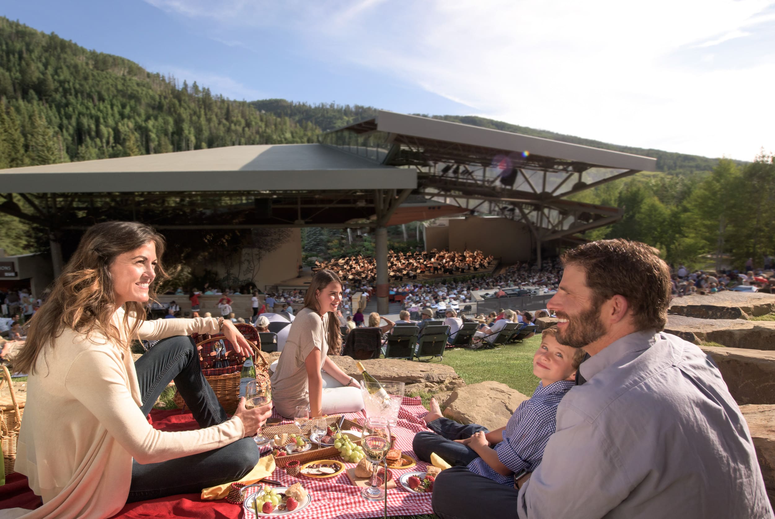 A family picnics on the lawn of The Amp in Vail while a Bravo! concert plays from the outdoor amphitheater. The sky is clear and blue overhead.