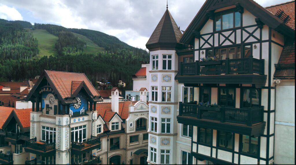European-style buildings stand tall and line the scenic streets in the Vail villages. Behind the buildings, tall mountains rise up covered in green grass and trees.