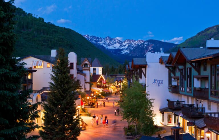 A streetlamp-lit street in the mountain village at dusk in Vail. People walk through the street between the clean, white-and-brown buildings and tall, verdant trees.
