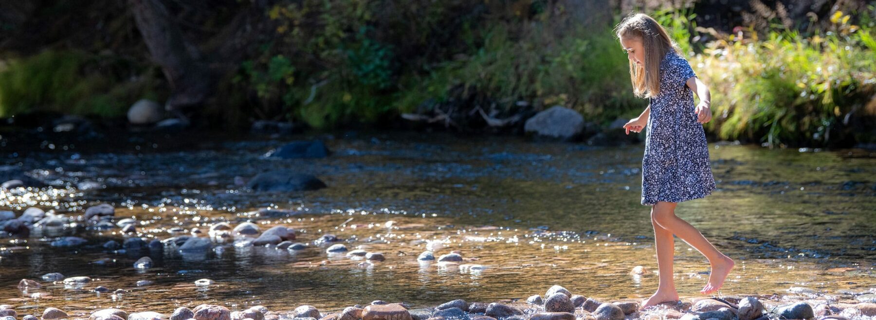 A girl wades in a shallow part of Gore Creek in Vail on a sunny summer day. She is barefoot in the water.