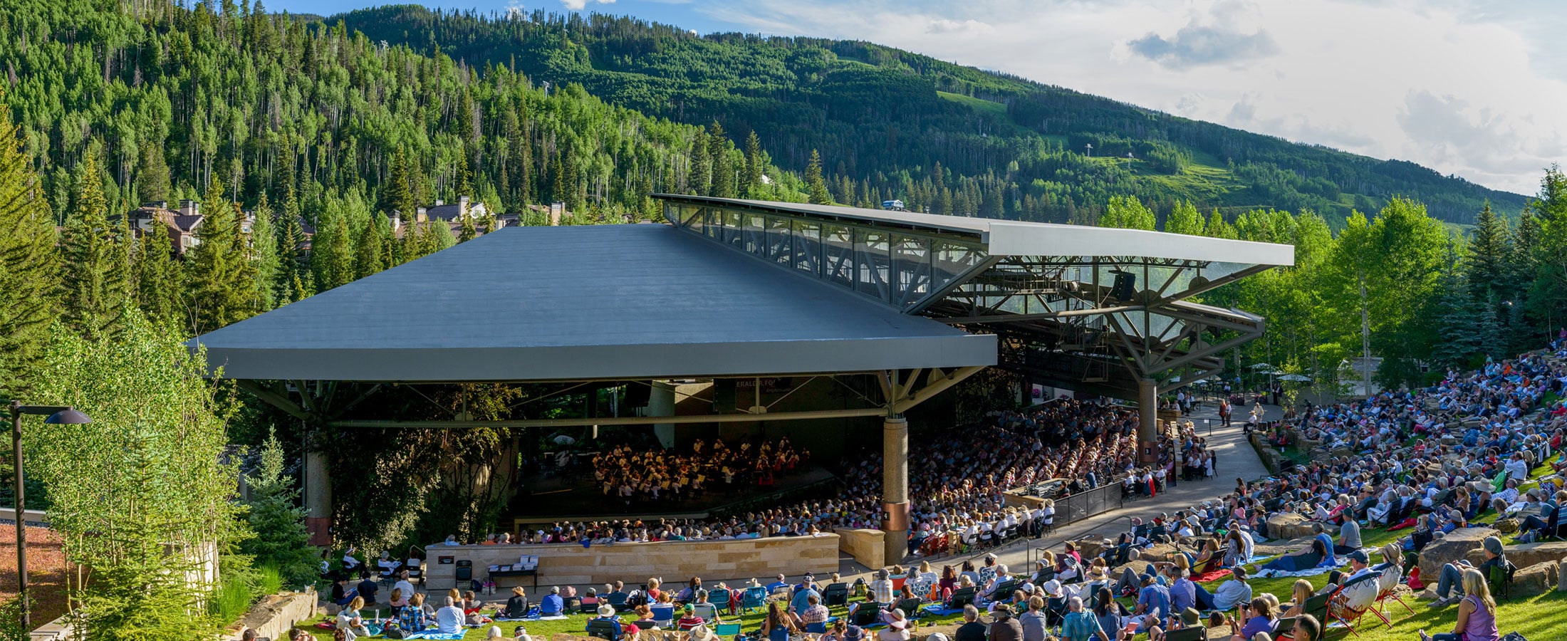Event attendees sit on blankets under a clear, blue sky and watch a performance at The Amp, an outdoor amphitheater in Vail. Evergreens stand on the rolling mountainside behind it.