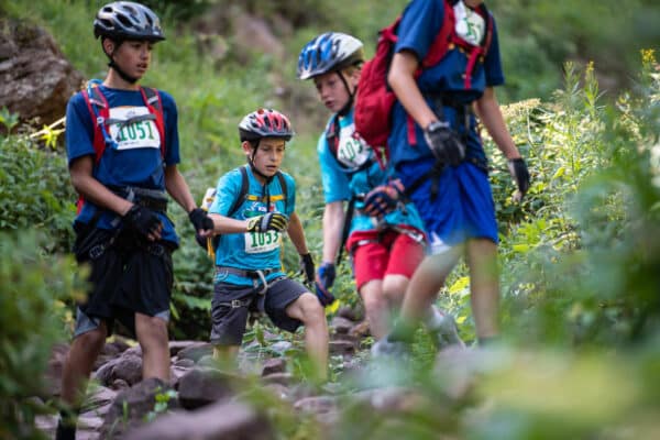 Four children wearing helmets and numbered bibs perform in a competition at Vail's Kids Adventure Games. They run over rocks and are surrounded by greenery.