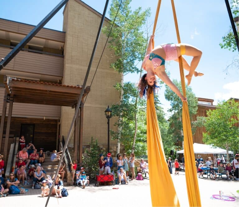 A person upside down, doing an aerial act in a yellow silk ribbon in front of a crowd during the Fourth of July celebration in Vail.
