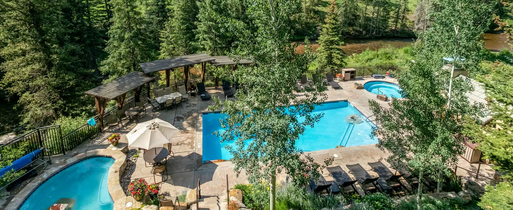 A turquoise-colored outdoor swimming pool at Antlers at Vail is surrounded by trees covered in green leaves, with mountains and a blue sky above.