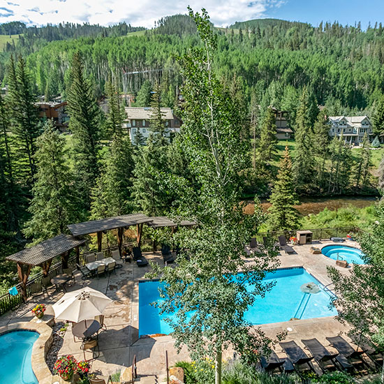 A turquoise-colored outdoor swimming pool at Antlers at Vail is surrounded by trees covered in green leaves, with mountains and a blue sky above.