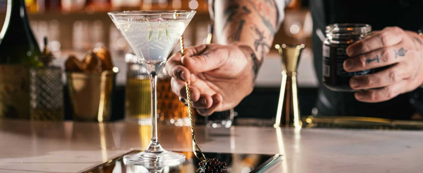 A bartender presents a martini on the bar of Chasing Rabbits in Vail. His tattooed arm reaches toward the frosty martini glass with bottles in the background.