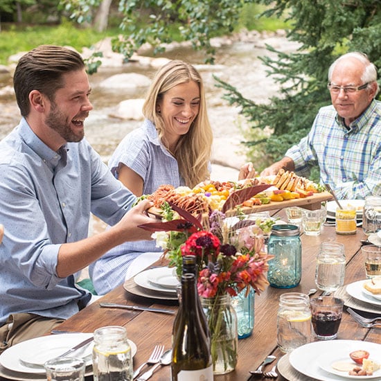 Five people gather around a table placed outdoors in Vail, CO, and dine on colorful plates of food with the rushing Gore Creek behind them.