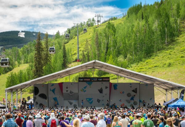 A GoPro Mountain Games climbing competition is watched by a large crowd with Vail Mountain's grassy, green ski slopes and gondola behind it.