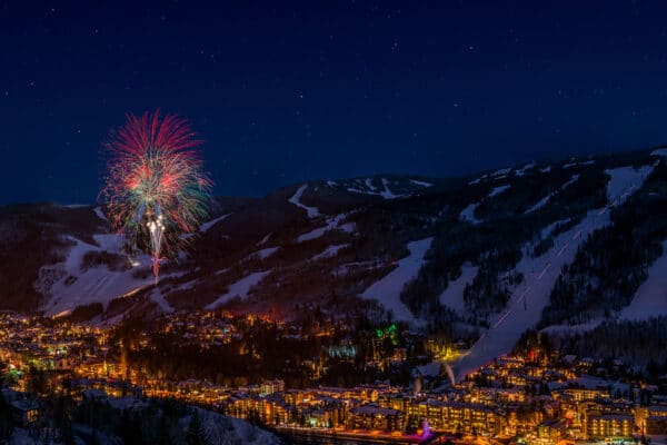 Fireworks burst over Vail mountain and the nearby villages. The mountain is covered in show and the villages are lit with lights.