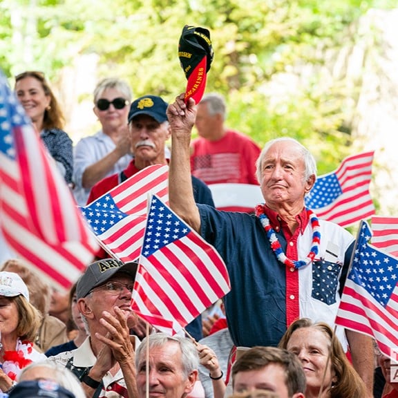 A man standing waving hat with american flags surrounding