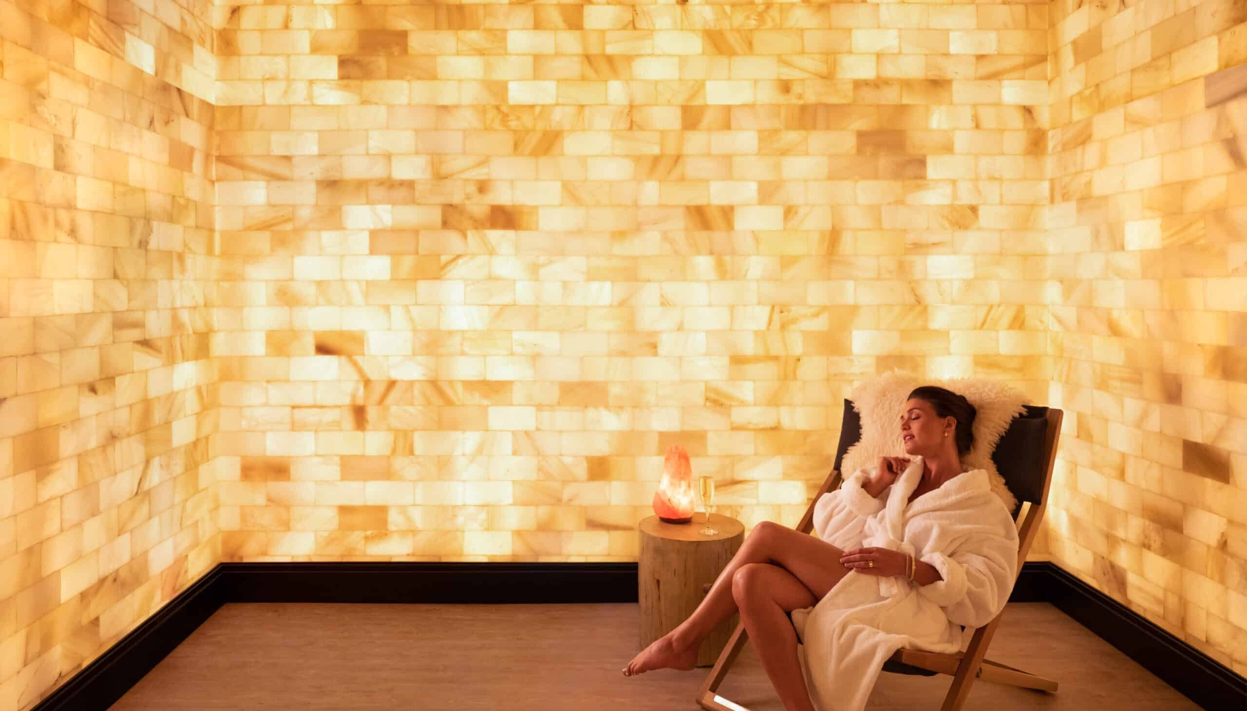 Woman at spa relaxing on chair in a salt room, a glass of wine and salt lamp sit beside her on a table