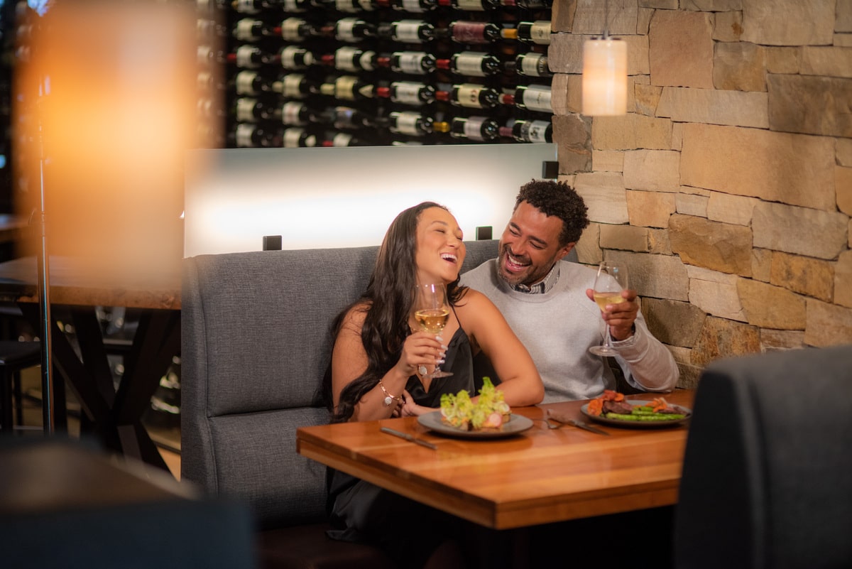 A couple laughs with glasses of white wine in their hands. They are seated beside each other in a cozy booth at Sweet Basil in Vail with beautifully plated dishes in front of them