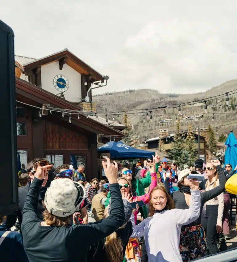 A crowd of people in winter clothing gather outdoors near a wooden building with a clock, raising their arms and enjoying a sunny day.