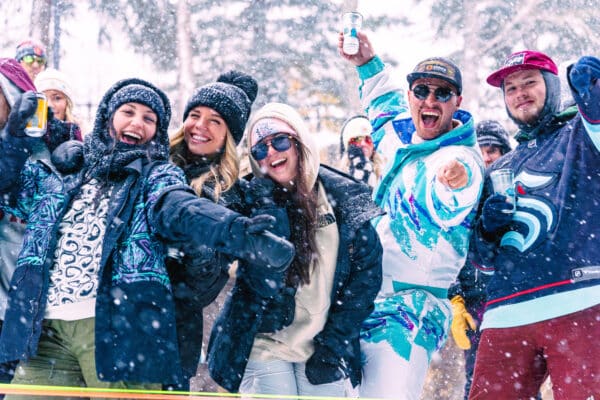 Group of five young people wearing snow gear pose for a photo in Vail, CO, and raise drinks up, arms out. They are smiling and laughing.