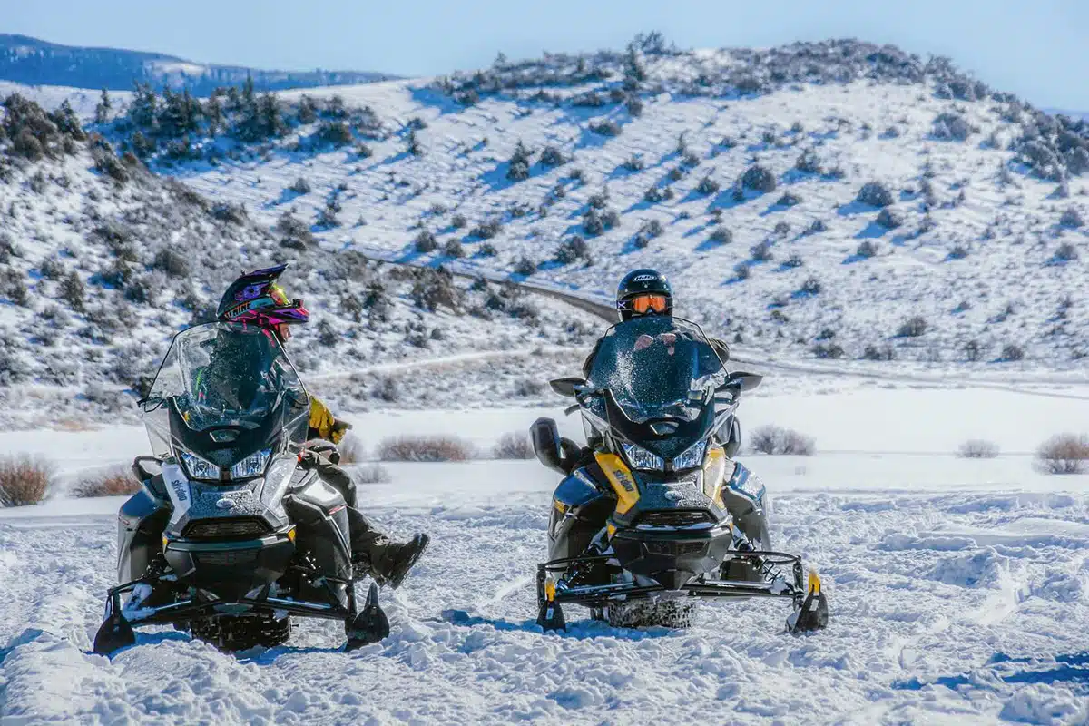 Two people sit on snowmobiles amid a snowy, alpine landscape near Vail, Colorado.