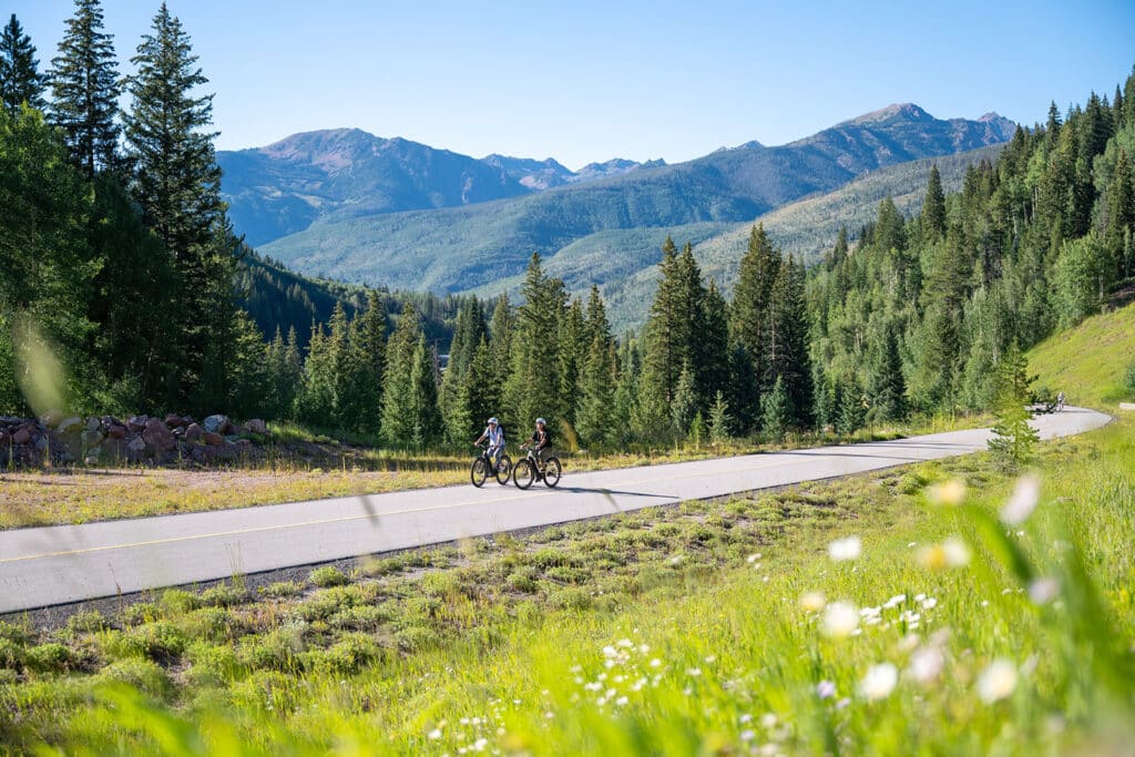 Two people e-biking on Vail Pass with mountains in the distance and wildflowers in the foreground
