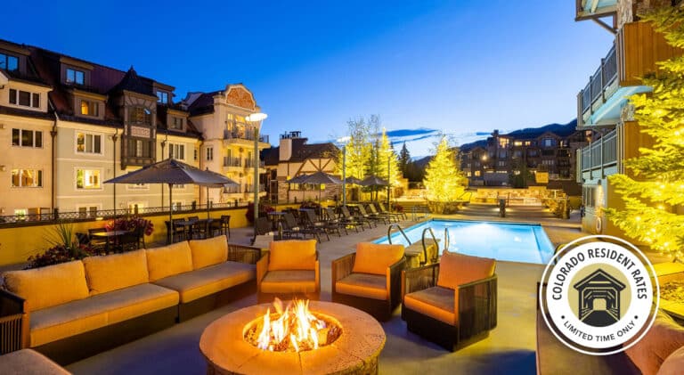 A fire pit outside the Landmark at Vail on a warm summer evening. The hotel pool and the nearby buildings can be seen nearby. A circle icon overlay in the bottom right reads "Colorado Resident Rates, Limited Time Only" with an illustration of a covered bridge.