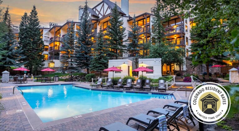 The pool deck outside Montaneros in Vail. The view from the corner of the pool shows the pool surface and surrounding walkways, chairs and umbrellas, and an illuminated luxury hotel building in the distance. A circle icon overlay in the bottom right reads "Colorado Resident Rates, Limited Time Only" with an illustration of a covered bridge.