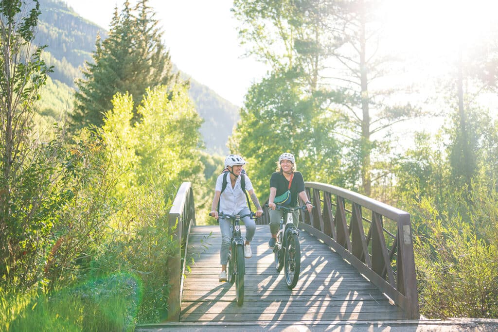Two people smile and e-bike across a wooden foot bridge in the mountain near Vail, Colorado.