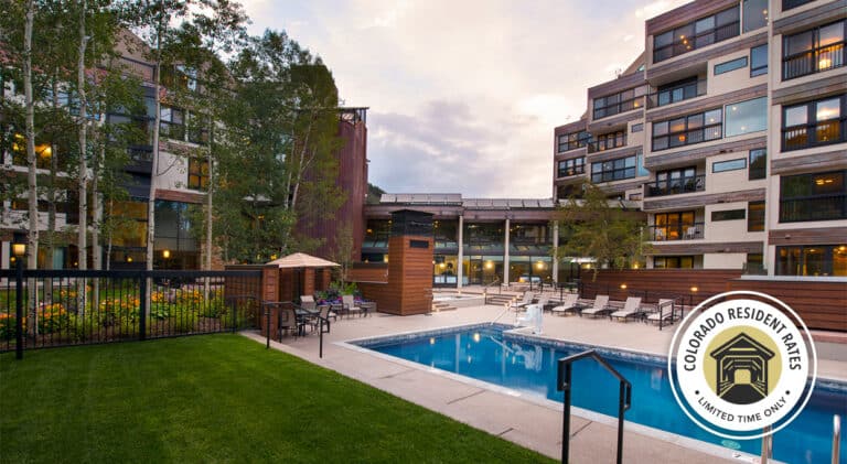 Exterior shot of the courtyard at Vail Spa. There is a grassy area and a pool in the foreground with several stories of rooms towering in the background. A circle icon overlay in the bottom right reads "Colorado Resident Rates, Limited Time Only" with an illustration of a covered bridge.