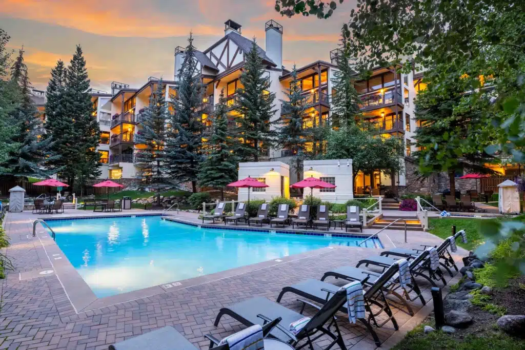 The pool deck outside Montaneros in Vail. The view from the corner of the pool shows the pool surface and surrounding walkways, chairs and umbrellas, and an illuminated luxury hotel building in the distance.