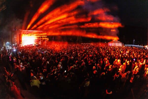 A large crowd attends an outdoor concert at night, illuminated by red stage lights and colorful spotlights, with a tent and trees visible in the background.