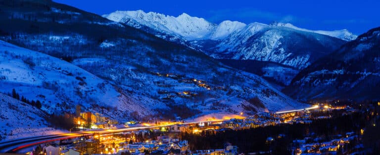 The snow-covered peaks surrounding Vail, Colorado, glow blue and white as evening falls as the villages are lit with warm lights.