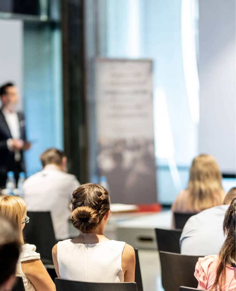 A view from the back of a room where a corporate workshop is happening. A man is presenting in the distance and a group of people are sitting intently or taking notes.