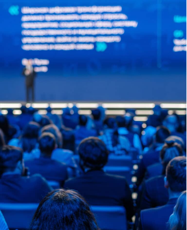 A keynote speaker gives a large on-screen presentation in a dimly lit and blue shaded auditorium.