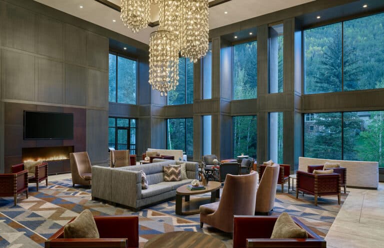 An upscale hotel lobby in Vail, Colorado with a decorative chandelier, fireplace, and a view of pine trees out the window.