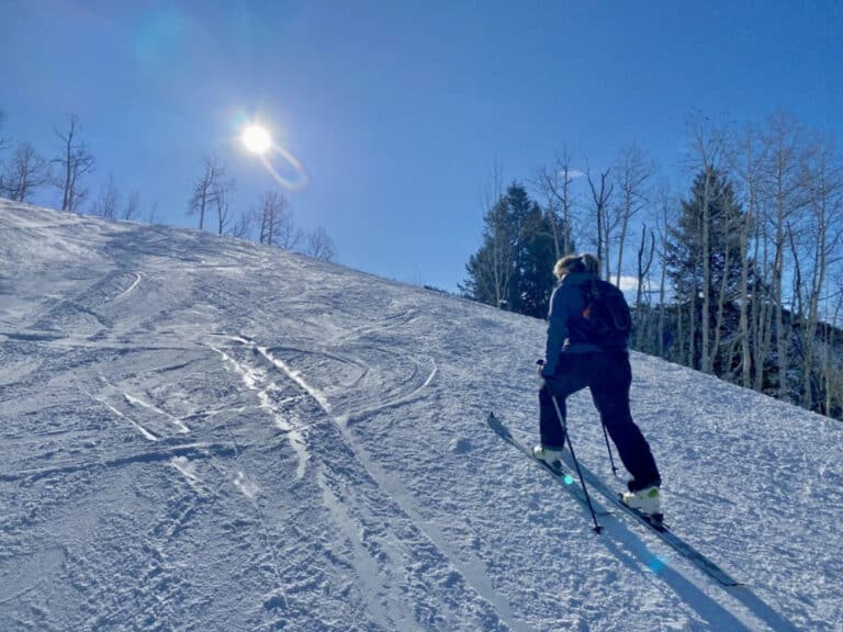 On Vail Mountain in Colorado, an uphill skier treks towards the top of top of a ski run with a bluebird sky, evergreens and bare aspen in the background.