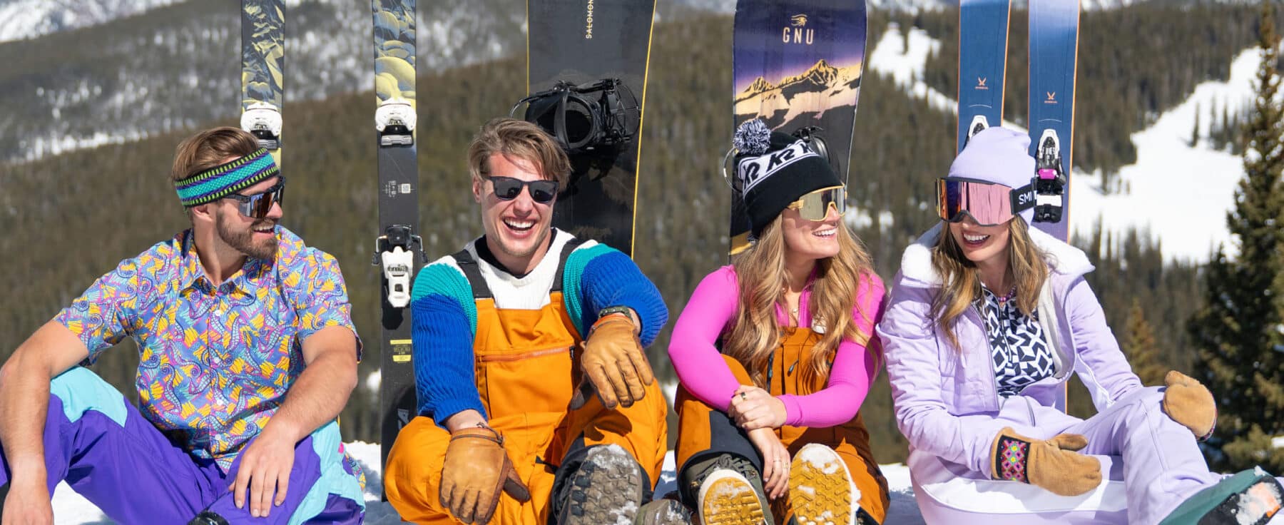 A group of four friends wearing colorful ski gear bask in the sun, laughing together, at Vail, Colorado.
