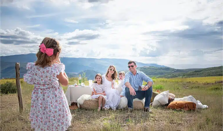 A family poses for a photo during an outdoor picnic with mountains in the background; a young girl with a pink bow faces them holding a camera.