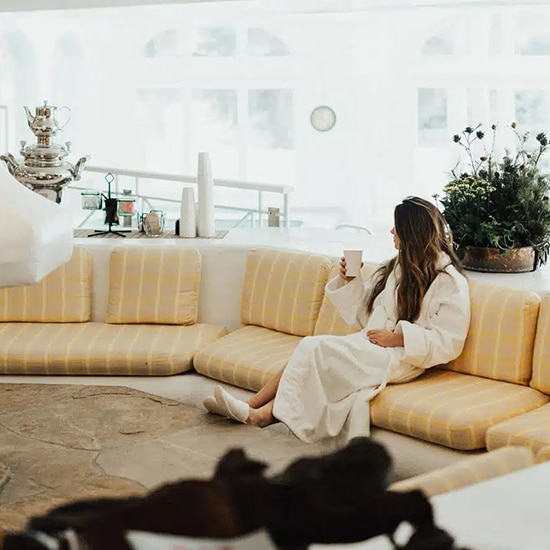 A spa guest sits in the Sonnenalp Spa relaxation room in Vail, Colorado, with a cup of tea. She wears a white, fluffy robe and gazes off in a contemplative fashion.